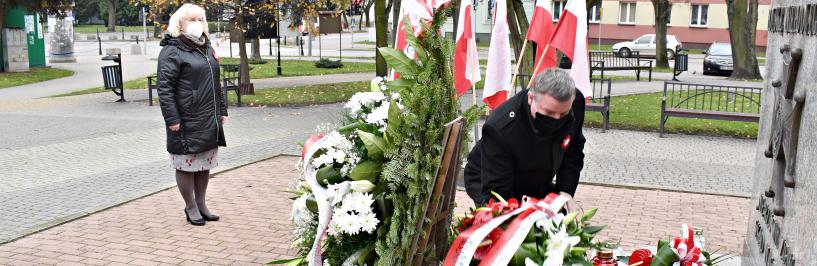 Burmistrz Jarosław Borowski składa wieniec biało-czerwonych kwiatów przed Pomnikiem Niepodległości Polski. Za nim stoi Zastępca Burmistrza. W tle biało-czerwone flagi.