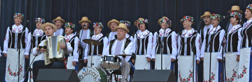 Na zdjęciu: członkowie zespołu Wasiloczki w białoruskich strojach ludowych na scenie bielskiego amfiteatru. Przed śpiewającymi dwaj mężczyźni: jeden grający na zestawie perkusyjnym, drugi grający na harmonii.