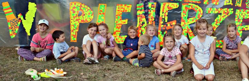 Grupa dzieci siedzących na trawie na tle przezroczystego baneru z kolorowym napisem o treści "czytelnia w plenerze"