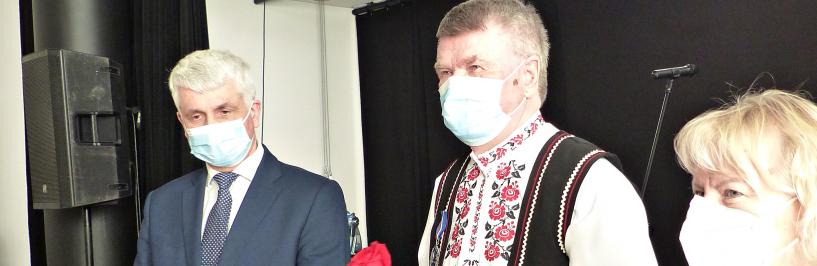 Ilustracja przedstawia trzy osoby stojące w maseczkach przed sceną domu kultury. W środku nagrodzony Sergiusz Łukaszuk, ubrany w strój ludowy i trzymający czerwoną różę. Po jego lewej stronie wojewoda, a po prawej stronie - zastępczyni burmistrza.