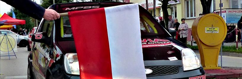 Na zdjęciu: samochód rajdowy na starcie imprezy za biało-czerwoną flagą startową, trzymaną przez burmistrza