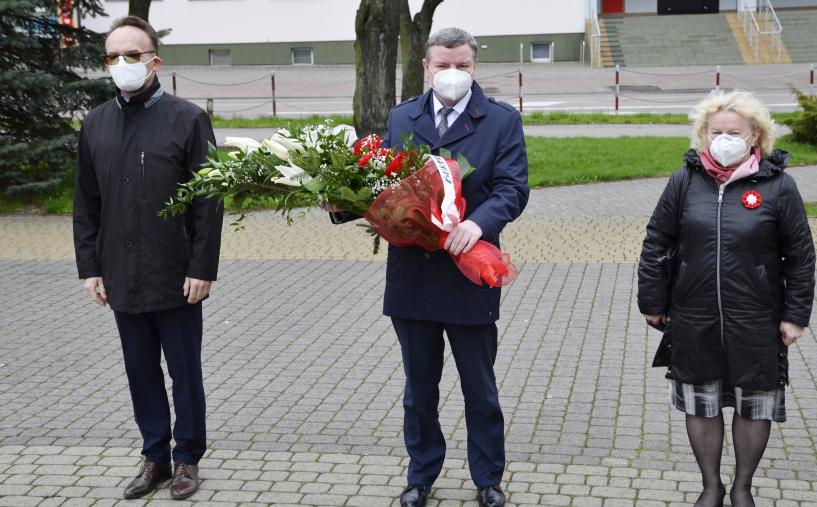 zdjęcie przedstawia trzy osoby: w środku burmistrz niosący wiązankę biało-czerwonych kwiatów, po prawej wiceburmistrz, po prawej wicestarosta