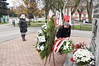 Burmistrz Jarosław Borowski składa wieniec biało-czerwonych kwiatów przed Pomnikiem Niepodległości Polski. Za nim stoi Zastępca Burmistrza. W tle biało-czerwone flagi.
