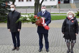 zdjęcie przedstawia trzy osoby: w środku burmistrz niosący wiązankę biało-czerwonych kwiatów, po prawej wiceburmistrz, po prawej wicestarosta