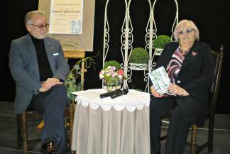 Ilustracja przedstawia dwie osoby siedzące na krzesłach przy stoliku przykrytym białym obrusem. Na stole stoi wazon z bukietem kwiatów i leżą dwa mikrofony. Po prawej stronie siedzi autorka książki Weronika Mielczarek, po lewej - prowadzący spotkanie Andrzej Salnikow.