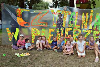 Grupa dzieci siedzących na trawie na tle przezroczystego baneru z kolorowym napisem o treści "czytelnia w plenerze"