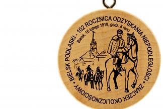 Ilustracja przedstawia okrągły drewniany znaczek turystyczny z wizerunkiem mężczyzn w mundurach, na koniach i z szablami, oraz ratuszem w tle. Wizerunek okala napis: "Bielsk Podlaski - 102. rocznica odzyskania niepodległości."